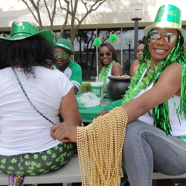 Four people in St. Patrick's Day attire, including green hats and beads, sitting outdoors.