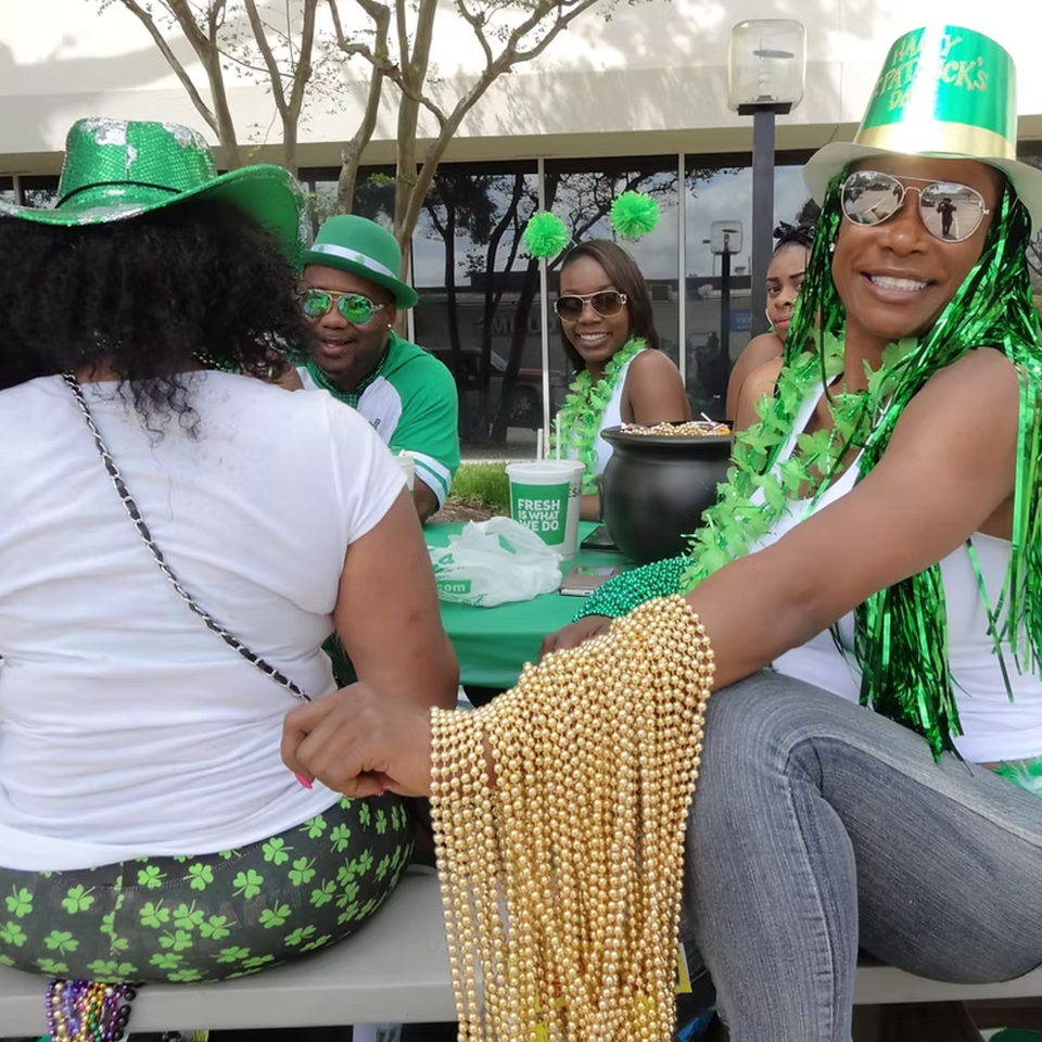Four people in St. Patrick's Day attire, including green hats and beads, sitting outdoors.