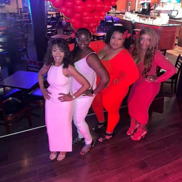 Four women posing in a bar-and-grill setting with red balloons and a bar in the background.