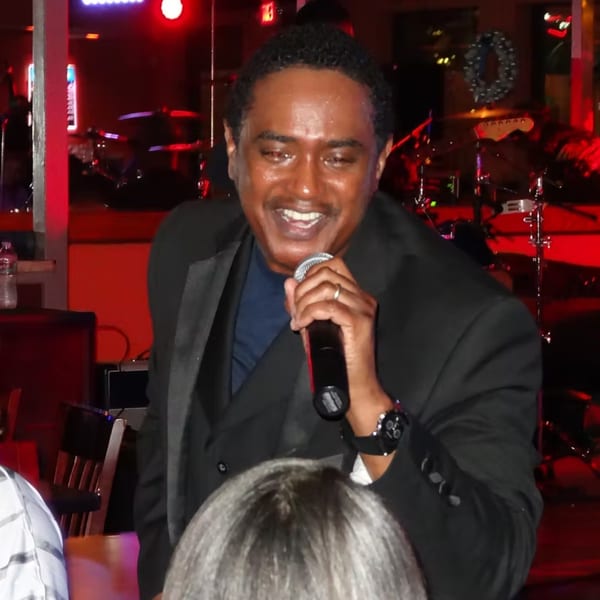 A man in a suit sings into a microphone on a dimly lit stage with red lighting.