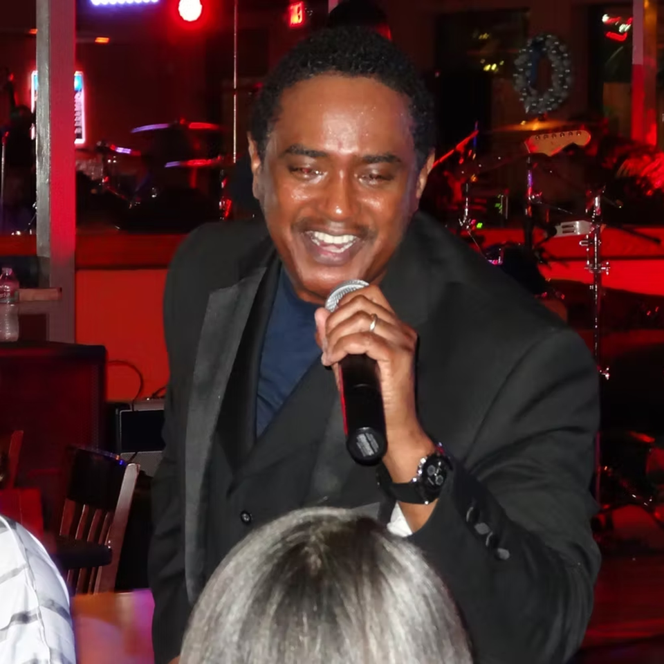 A man in a suit sings into a microphone on a dimly lit stage with red lighting.