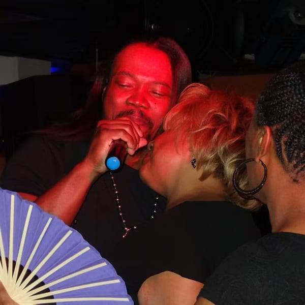 Man singing into a microphone, illuminated by red light, with two women beside him at Chicago Bar & Grill.