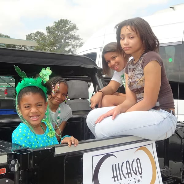Four young girls, some wearing green shamrock accessories, sitting in the back of a vehicle.