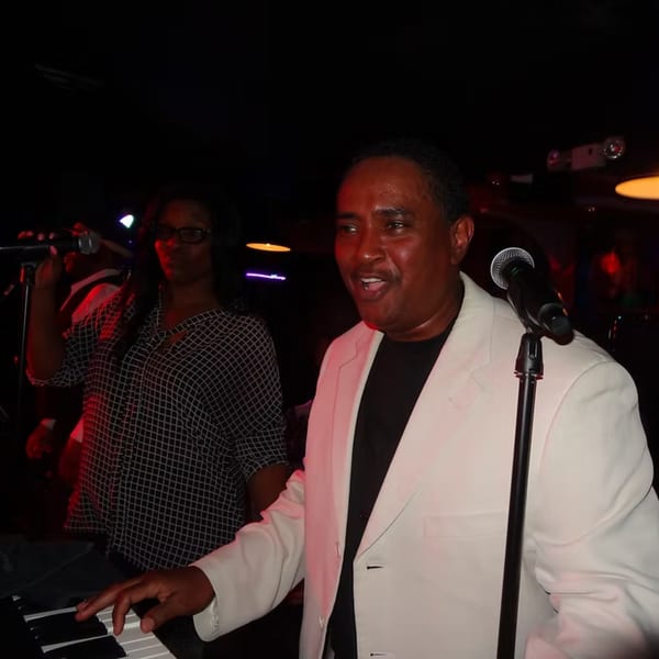 A man in a white jacket sings into a microphone while playing a keyboard, with a woman singing behind him.