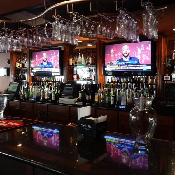A well-stocked bar at Chicago Bar & Grill with liquor bottles, hanging glasses, and two TVs showing sports.