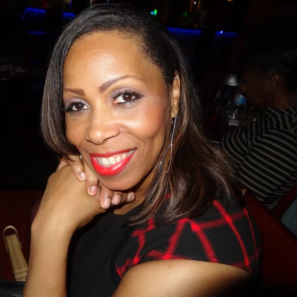 Smiling woman with dark hair and red lipstick, wearing a black and red plaid top, in a dimly lit bar.