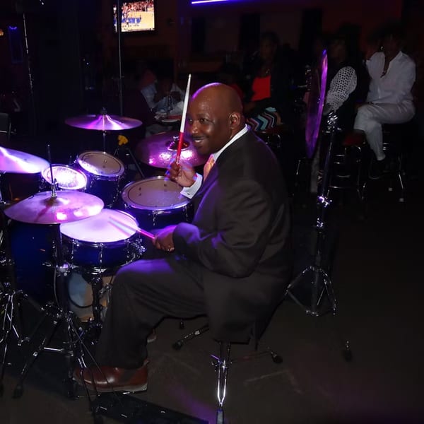 A smiling man in a suit plays drums under purple lights at Chicago Bar & Grill.