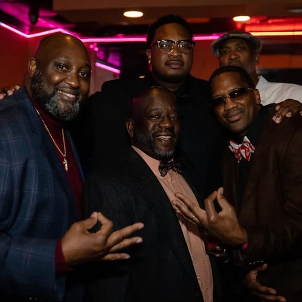 Five men in suits and bow ties posing together in a dimly lit bar with pink neon lights.