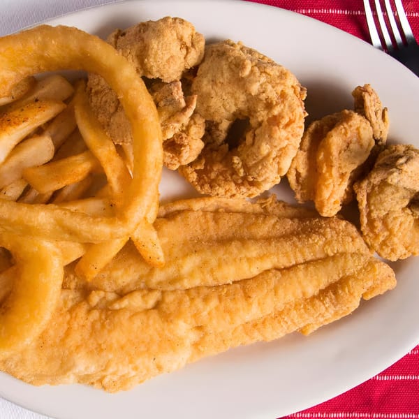 A white plate holds fried fish, shrimp, onion rings, and french fries on a red and white placemat.