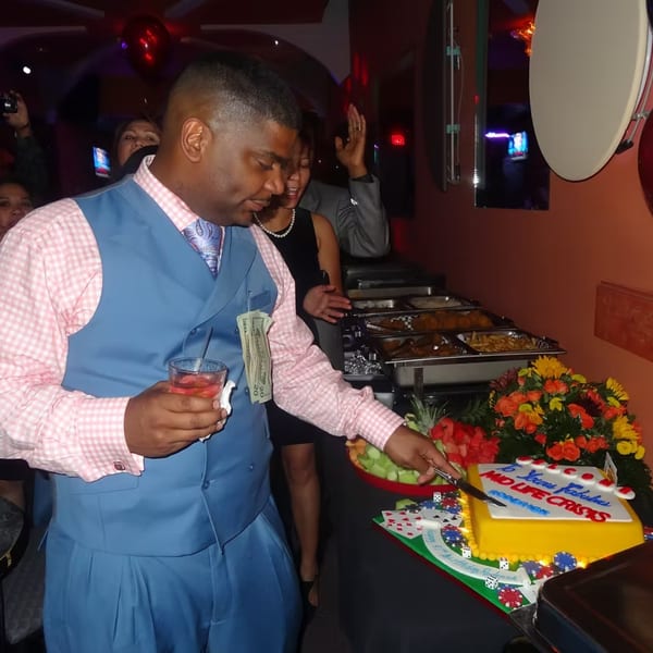 Man in blue vest and pink shirt cutting a yellow cake with "NO LIFE CRISIS" written on it.
