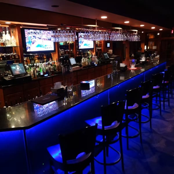 A long, dark bar with blue under-lighting, bar stools, liquor bottles, and TVs showing sports.