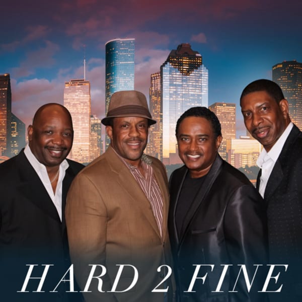 Four men in suits and a fedora smiling in front of a city skyline at dusk.